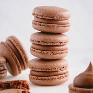 Three filled chocolate macarons stacked on top of each other surrounded by other chocolate macarons.