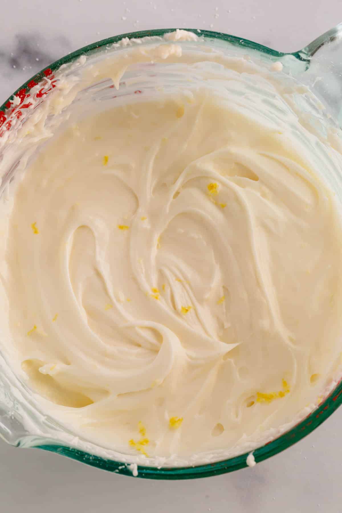 A liquid measuring cup full of lemon cream cheese frosting with flakes of lemon zest.