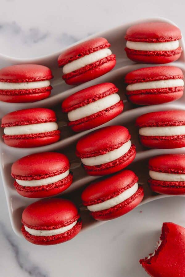 Vibrant red velvet macarons filled with white cream cheese frosting arranged on a white serving platter.
