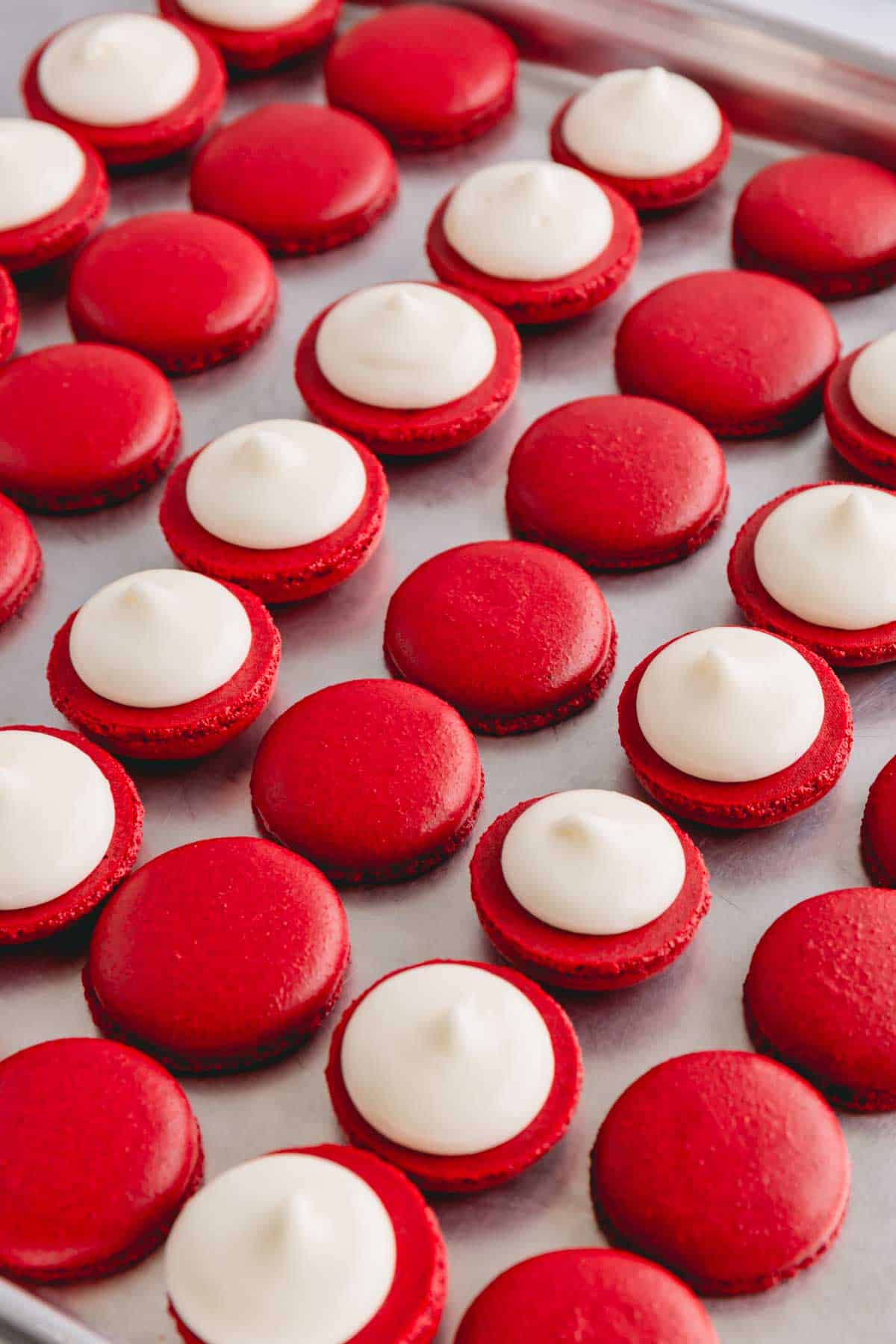Red velvet macaron shells arranged on a baking sheet and half of the shells with cream cheese frosting.