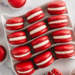 Vibrant red velvet macarons filled with white cream cheese frosting arranged on a white serving platter.