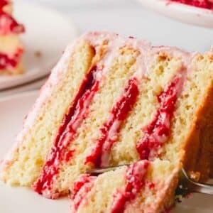 A slice of raspberry layer cake with raspberry jam on a plate with a fork cutting a bite.