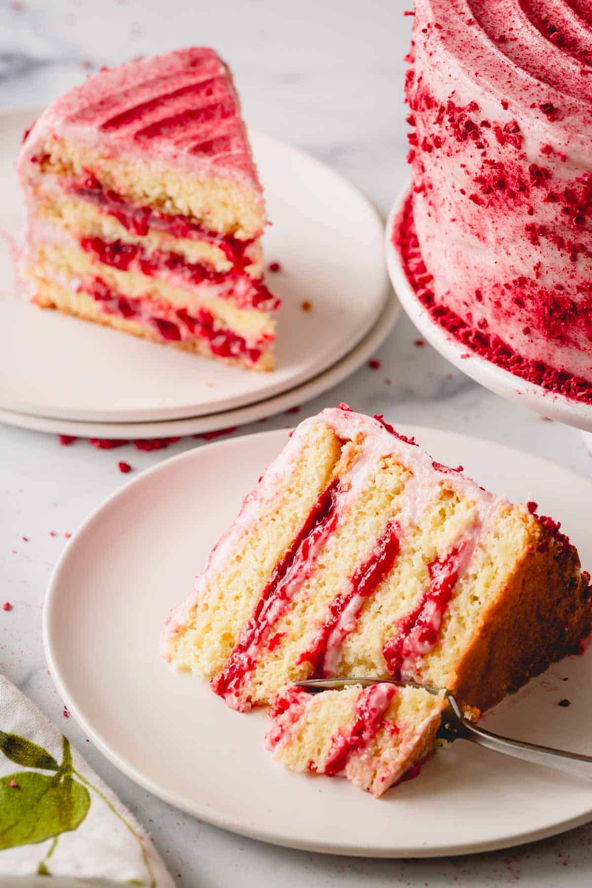 Two slices of raspberry layer cake on plates next to the whole cake and a fork cutting a bite of one.