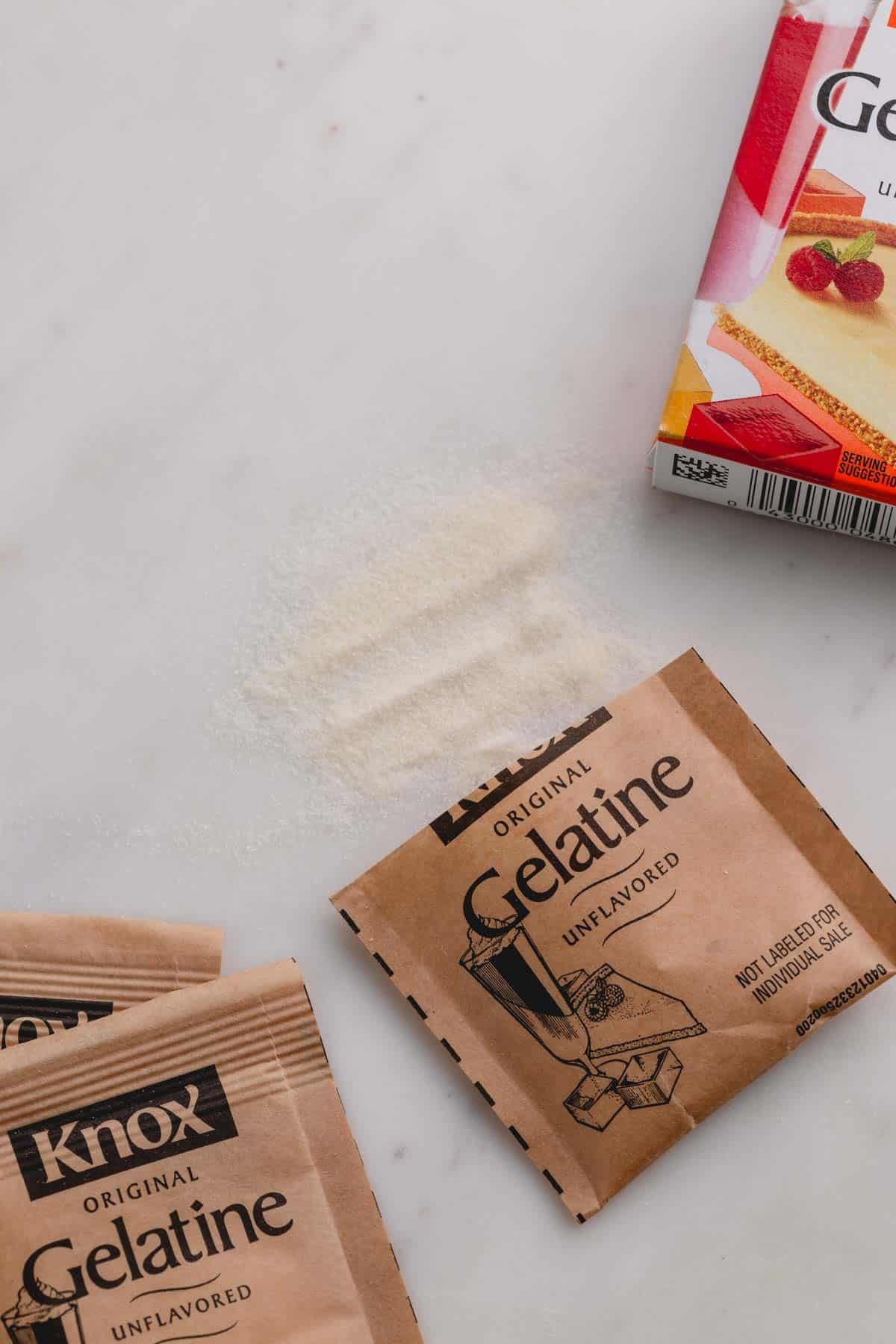 Packets of Knox unflavored gelatin on a white background and some gelatin powder spilled out.