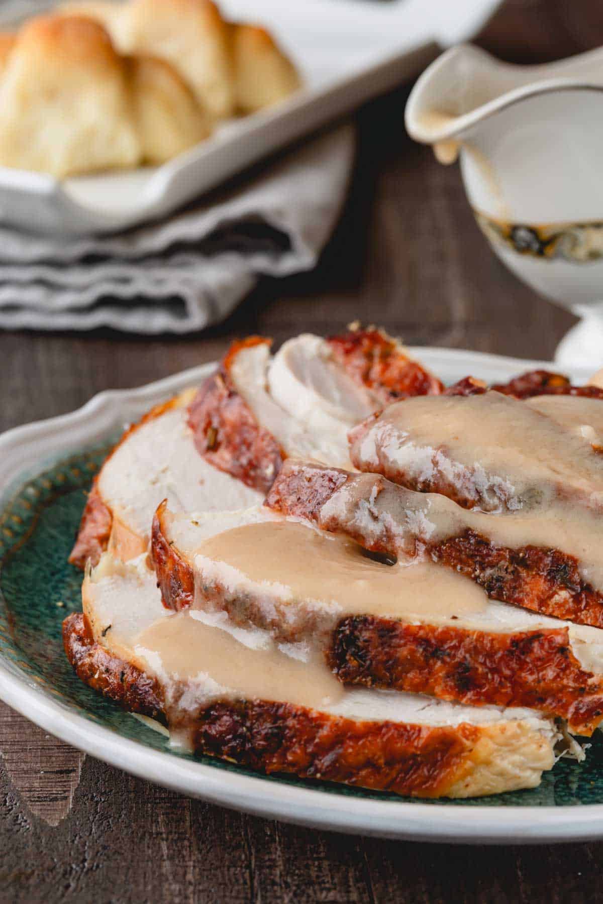 Turkey slices on a plate topped with gravy with the gravy boat and rolls in the background.