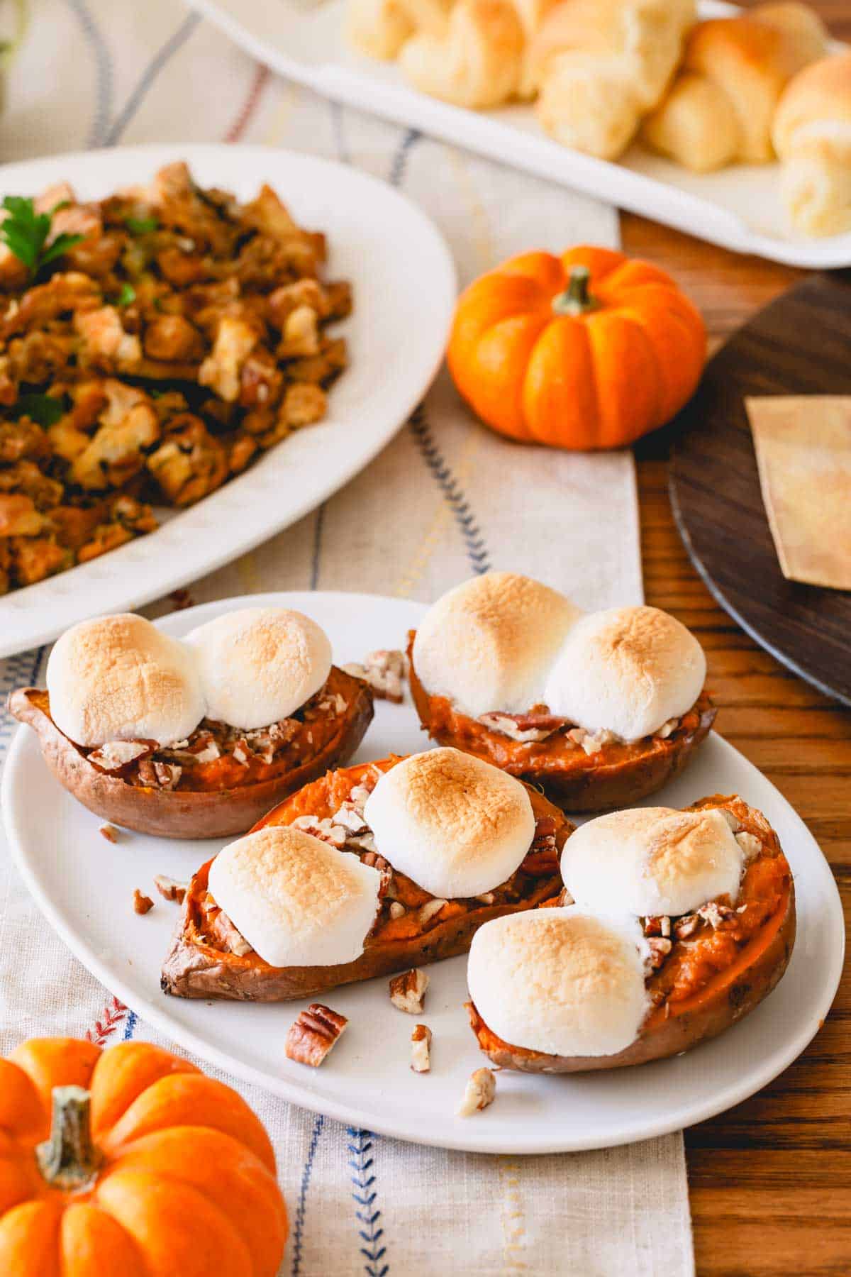 Toasted marshmallow twice baked sweet potatoes on a white plate along with other plates of stuffing and dinner rolls.