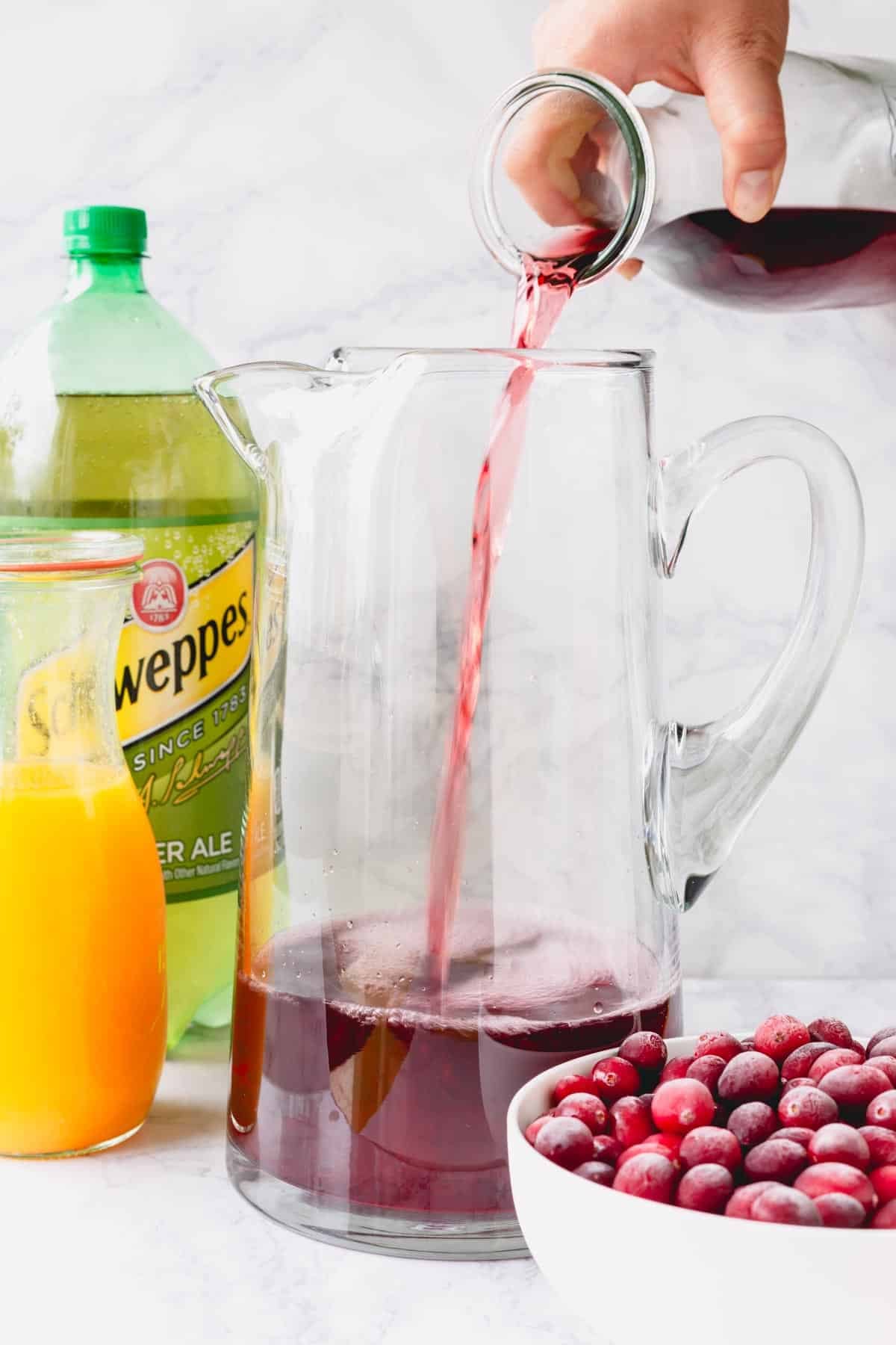 Cranberry juice being poured into a pitcher with ginger ale, orange juice, and pomegranates next to it.