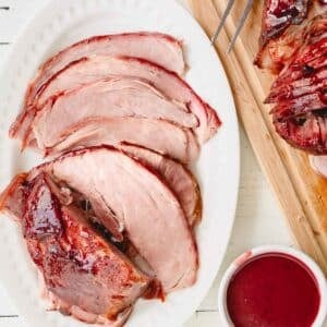 Overhead image of glazed spiral ham on a wooden cutting board and a serving platter with a bowl of glaze.