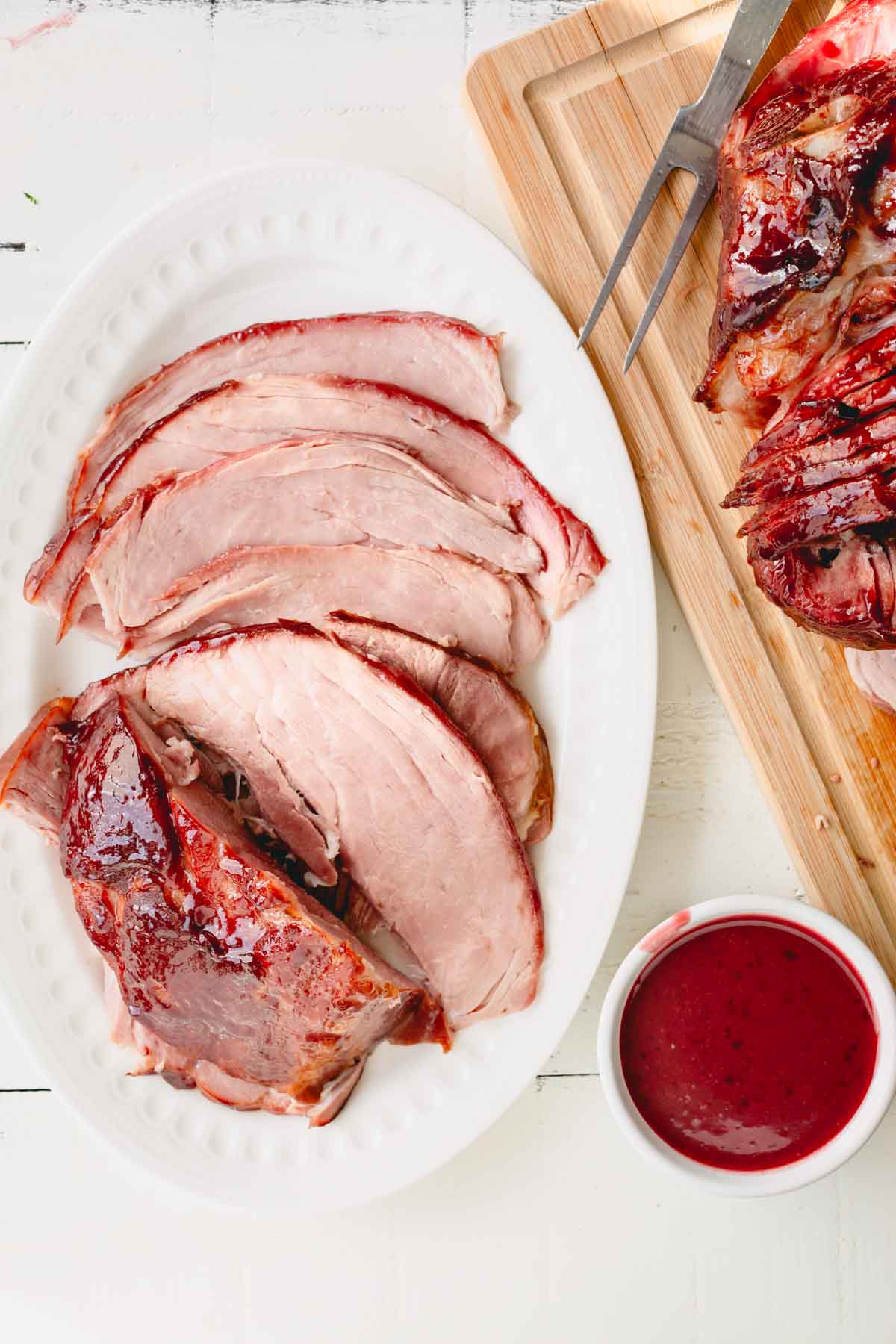 Overhead image of glazed spiral ham on a wooden cutting board and a serving platter with a bowl of glaze.