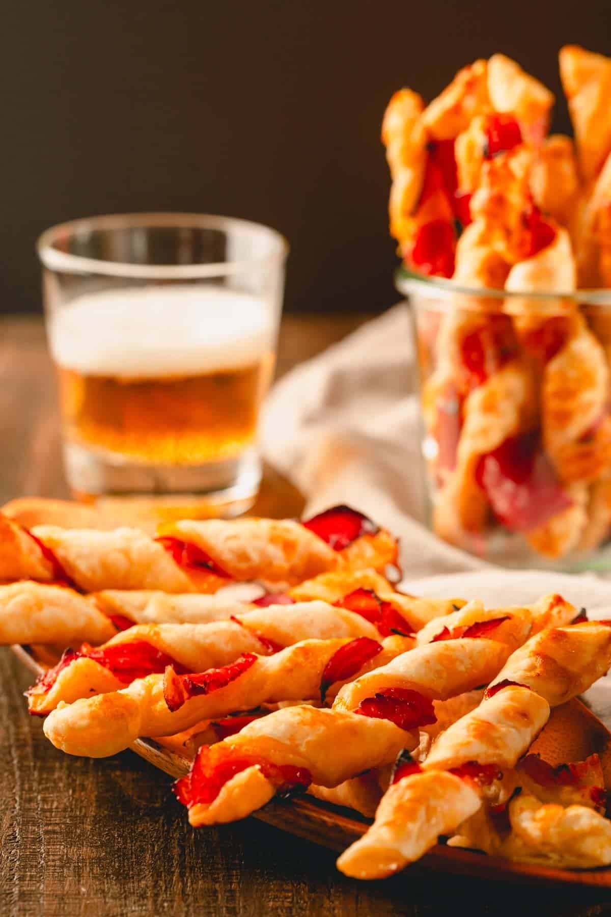 Prosciutto puff pastry twists on a plate and in a glass cup with a glass of beer in the background.