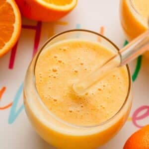 A glass of coconut orange Julius with a glass straw sticking out and oranges around it.