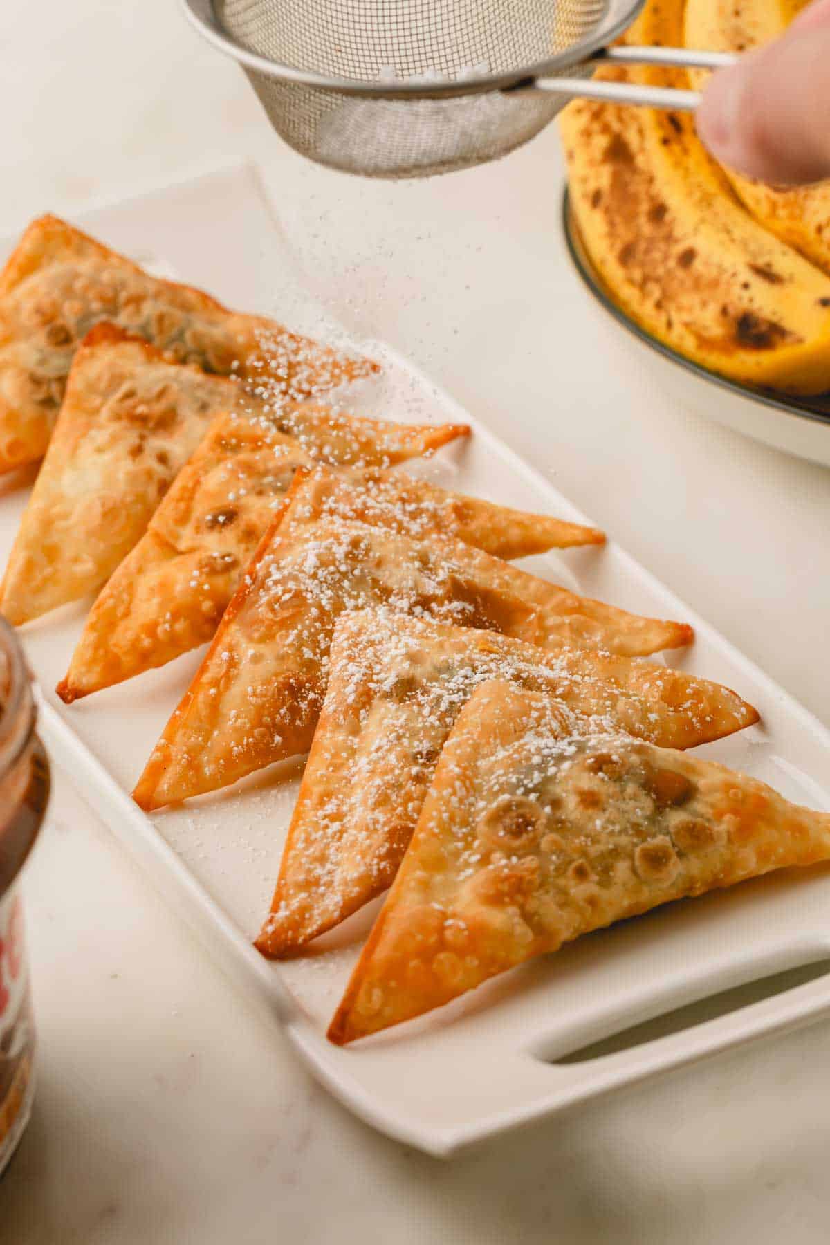 A platter of banana and Nutella wontons topped with powdered sugar being dusted over the top.