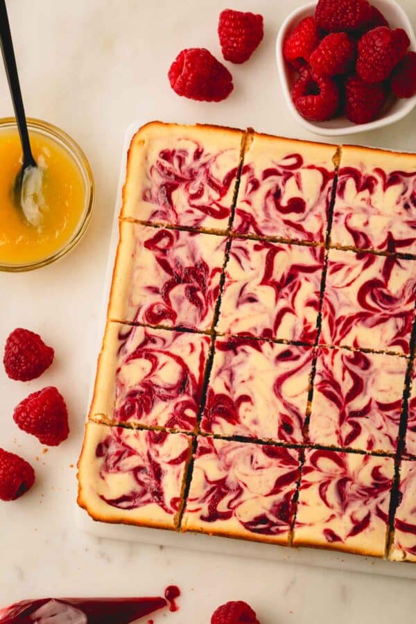 Lemon raspberry cheesecake bars next to fresh raspberries and a bowl of lemon curd.
