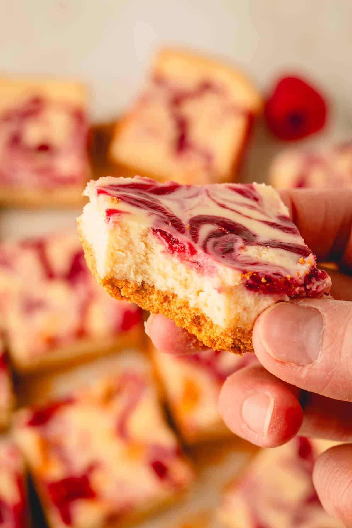 A hand holding a lemon raspberry cheesecake bar with a corner bite missing.