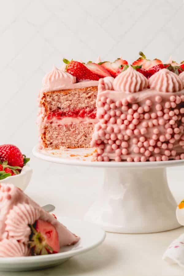 A two layer frosted strawberry cake on a cake stand with a slice missing.