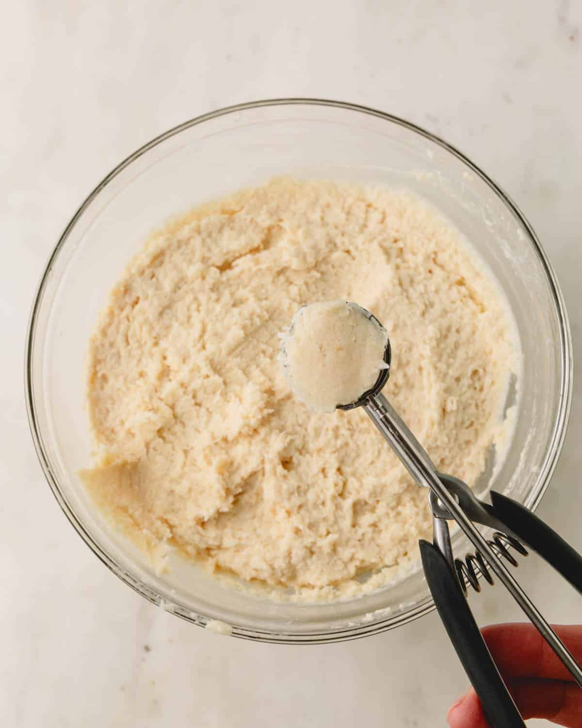 A bowl of almond coconut balls batter with a cookie scoop full of dough held above it.