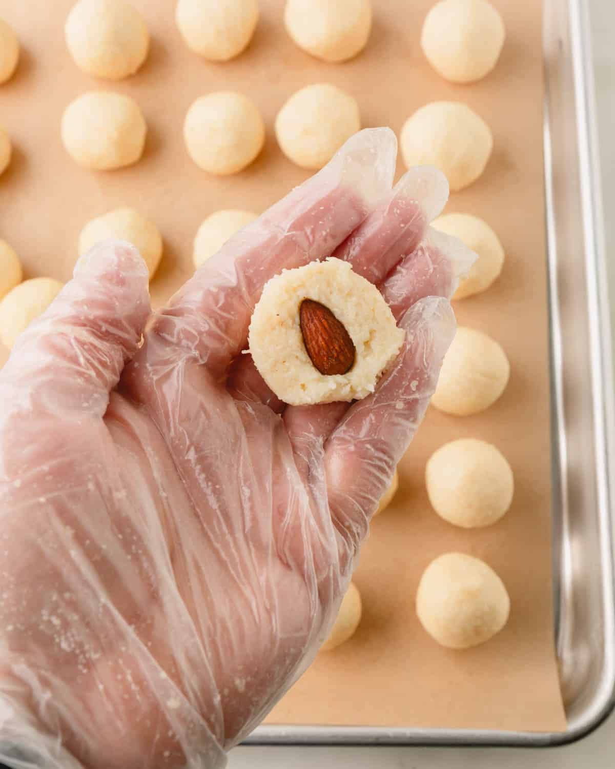 A hand holding a coconut ball with a hole pecan in the center above a sheet tray of almond coconut balls.