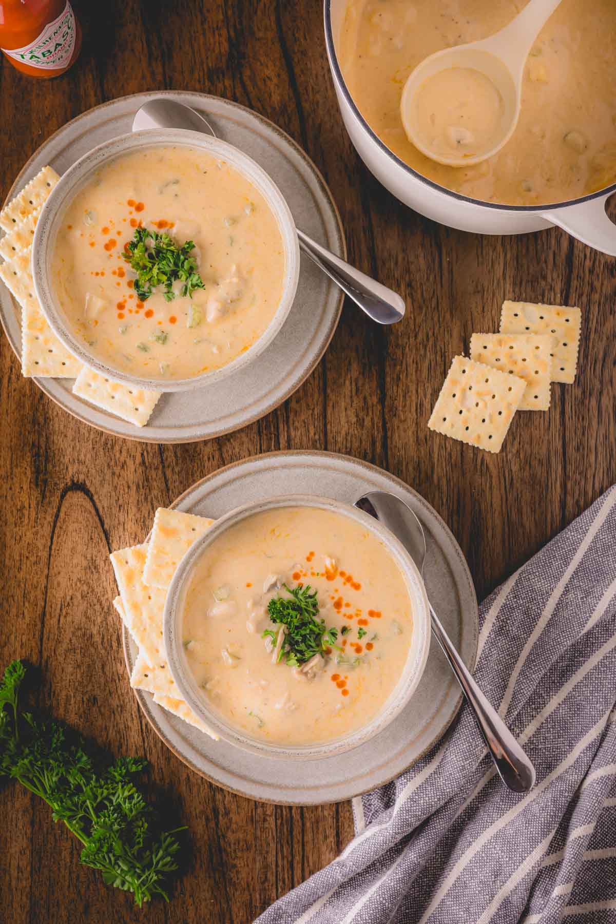 Two bowls of oyster stew on plates with crackers and spoons next to a pot of oyster stew.