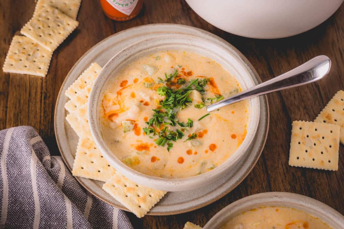 A bowl of oyster stew with a spoon sticking out on a plate surrounded by crackers.