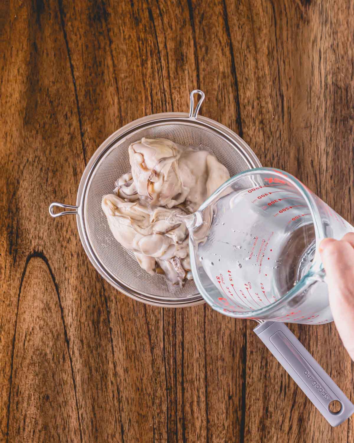 Oysters in a sieve over a bowl, water being poured on top.