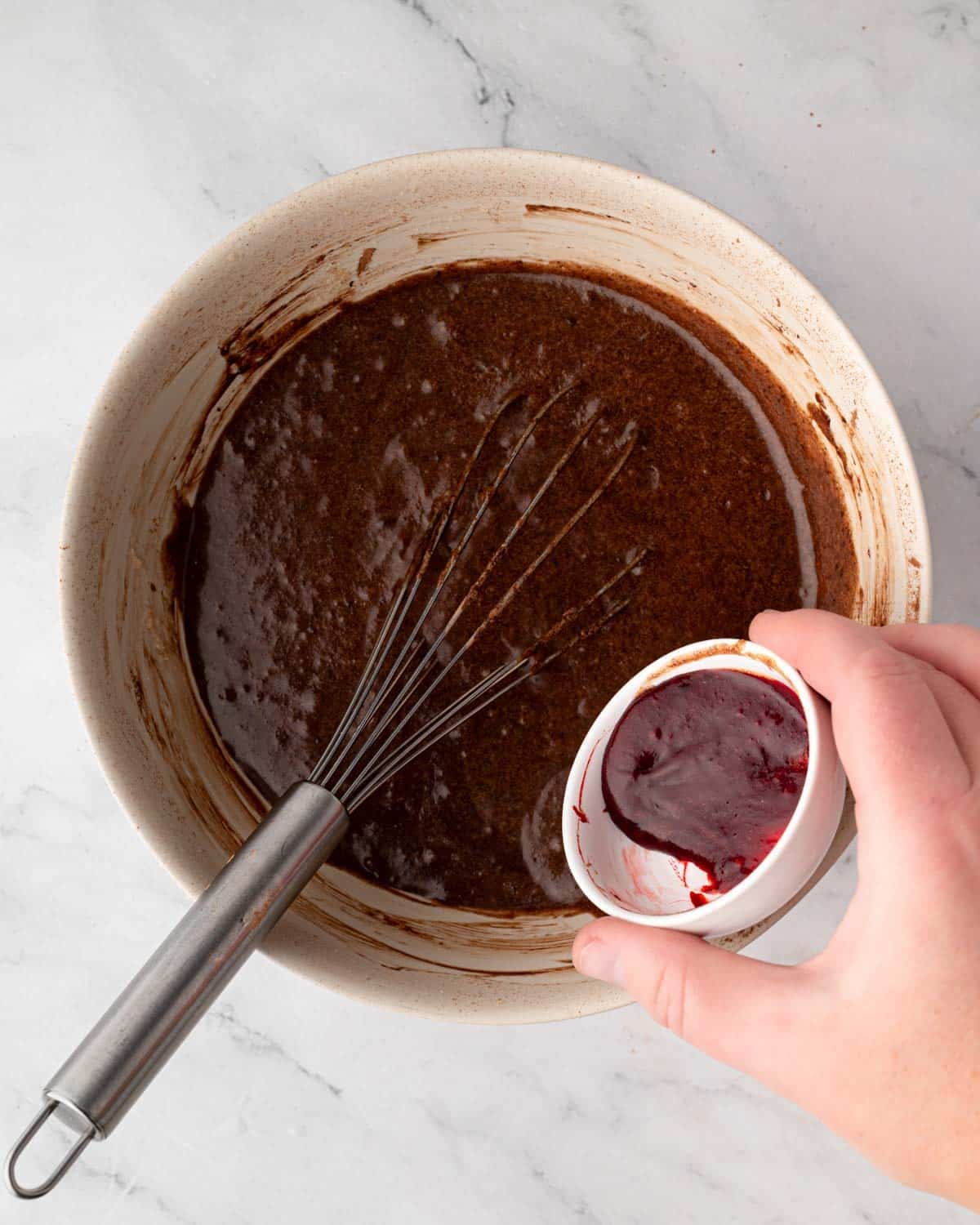 Red gel food coloring being added to a bowl of red velvet brownie batter, a whisk sticking out of the bowl.