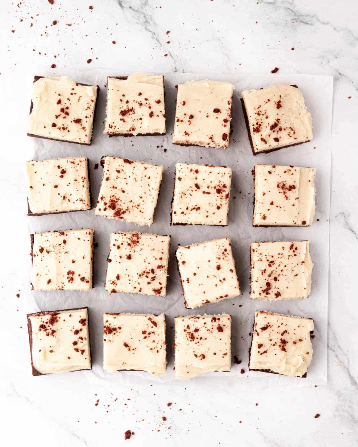 Red velvet brownie squares with cream cheese frosting on a sheet of parchment paper.