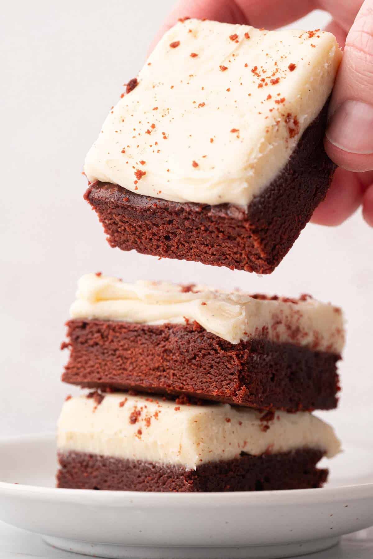 A stack of three red velvet brownies topped with cream cheese frosting, a hand lifting the top brownie up.
