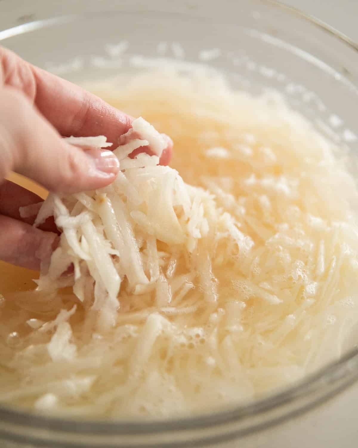 A hand soaking grated potatoes in a glass bowl in ice water.