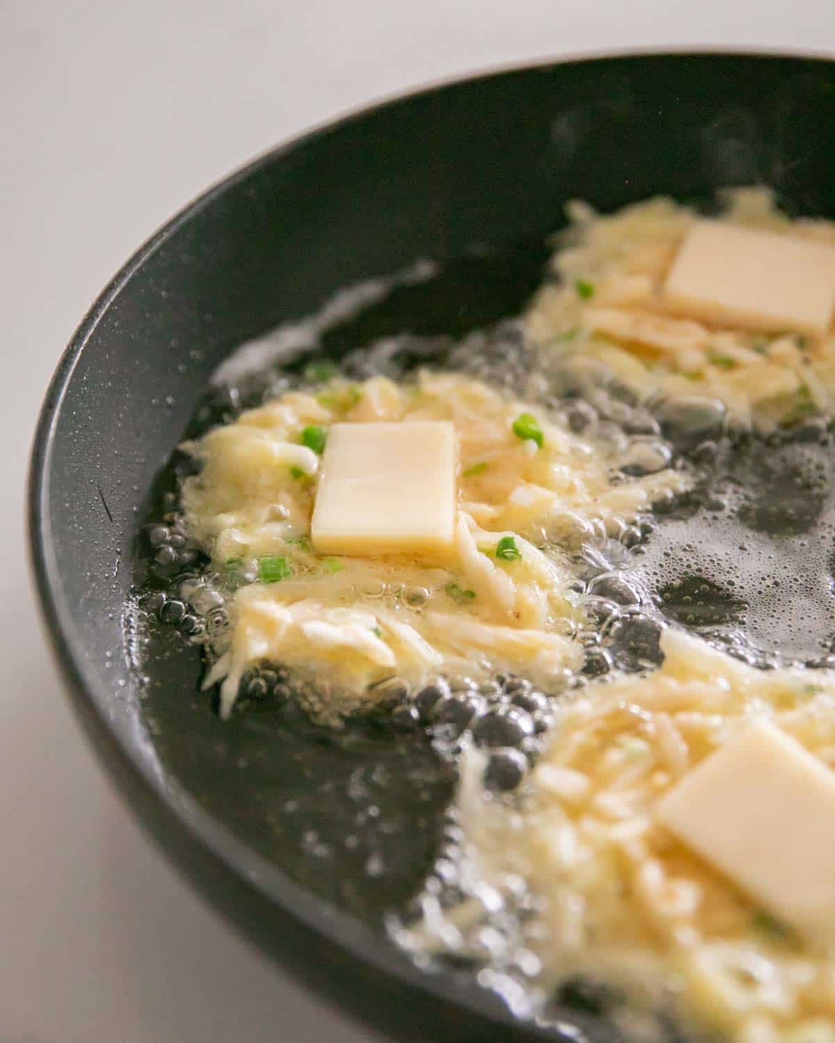 A large skillet with cheesy potato pancakes cooking and a slice of cheese in the center.