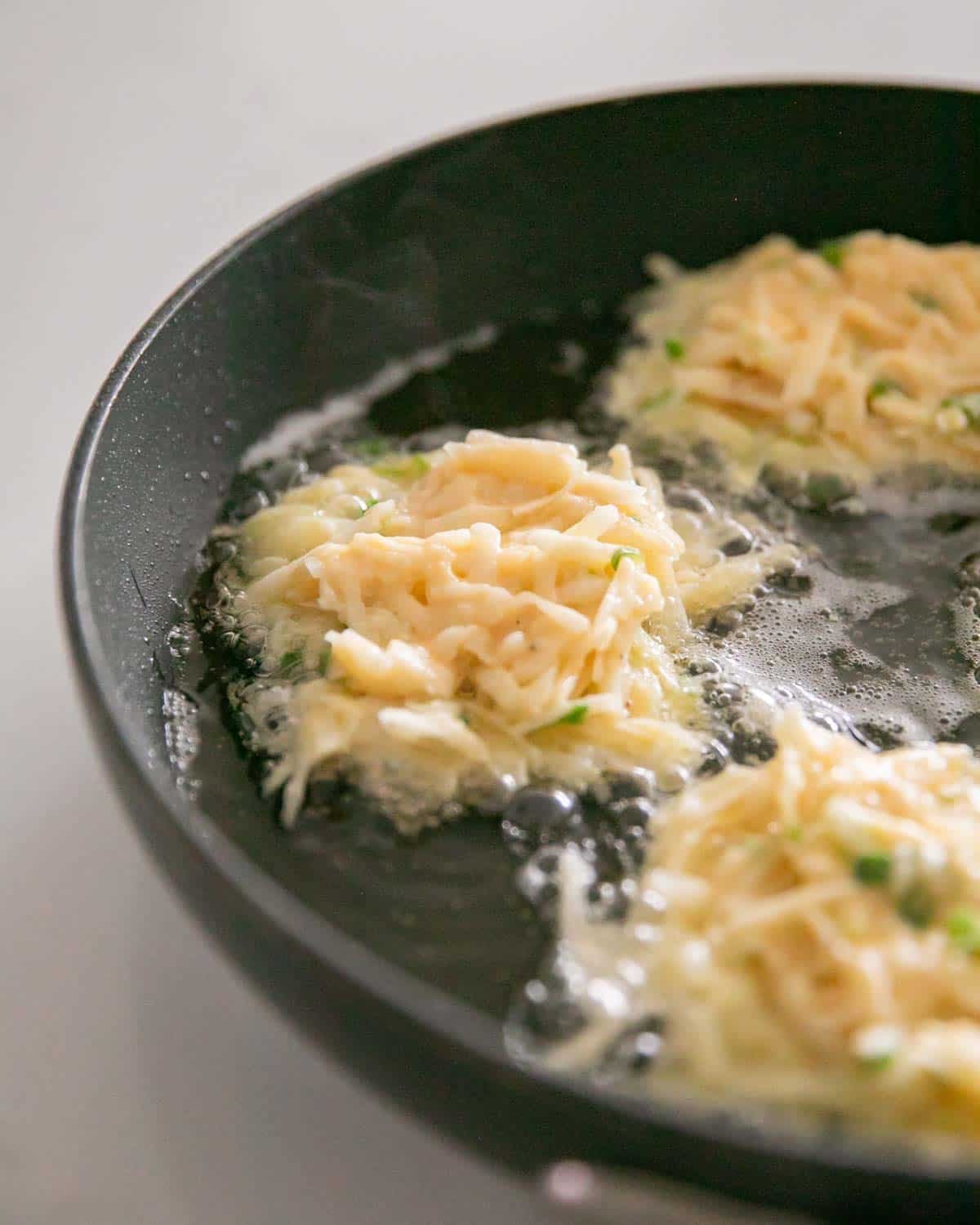 A skillet cooking cheesy potato pancakes with more grated potato on top.