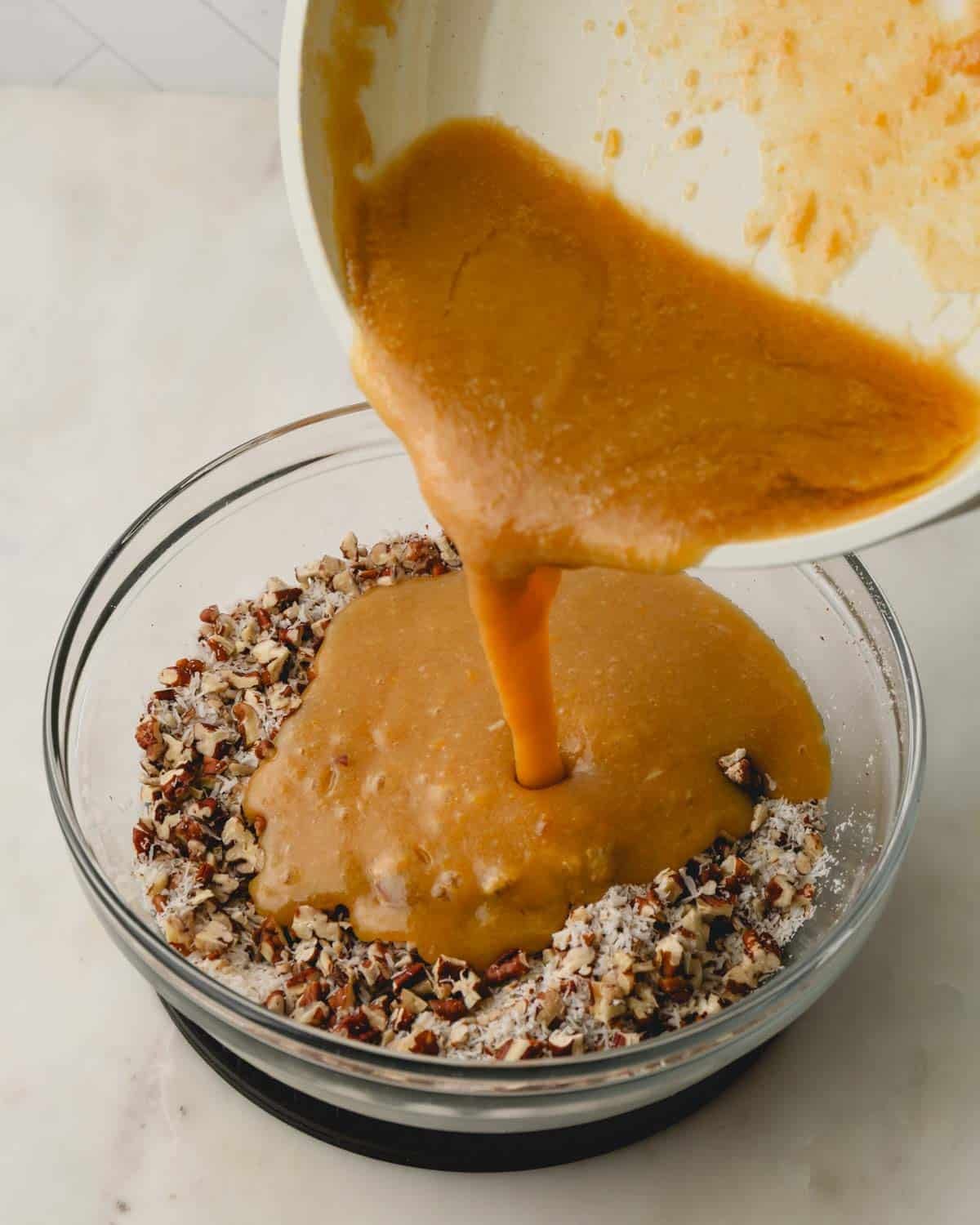 A pan with melted brown sugar and butter mixture being poured over coconut and pecans in a glass bowl.