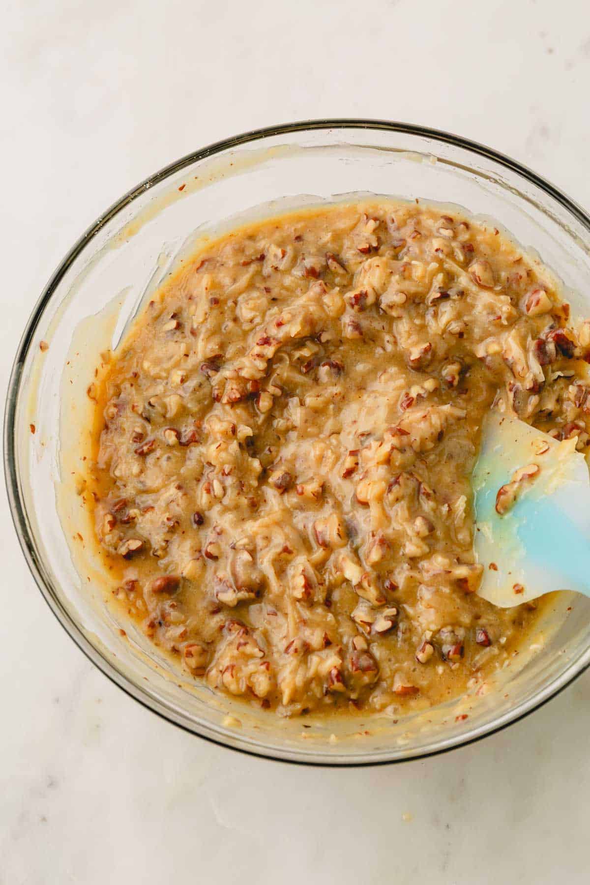 A glass bowl with coconut and pecans and a spatula resting on the side.
