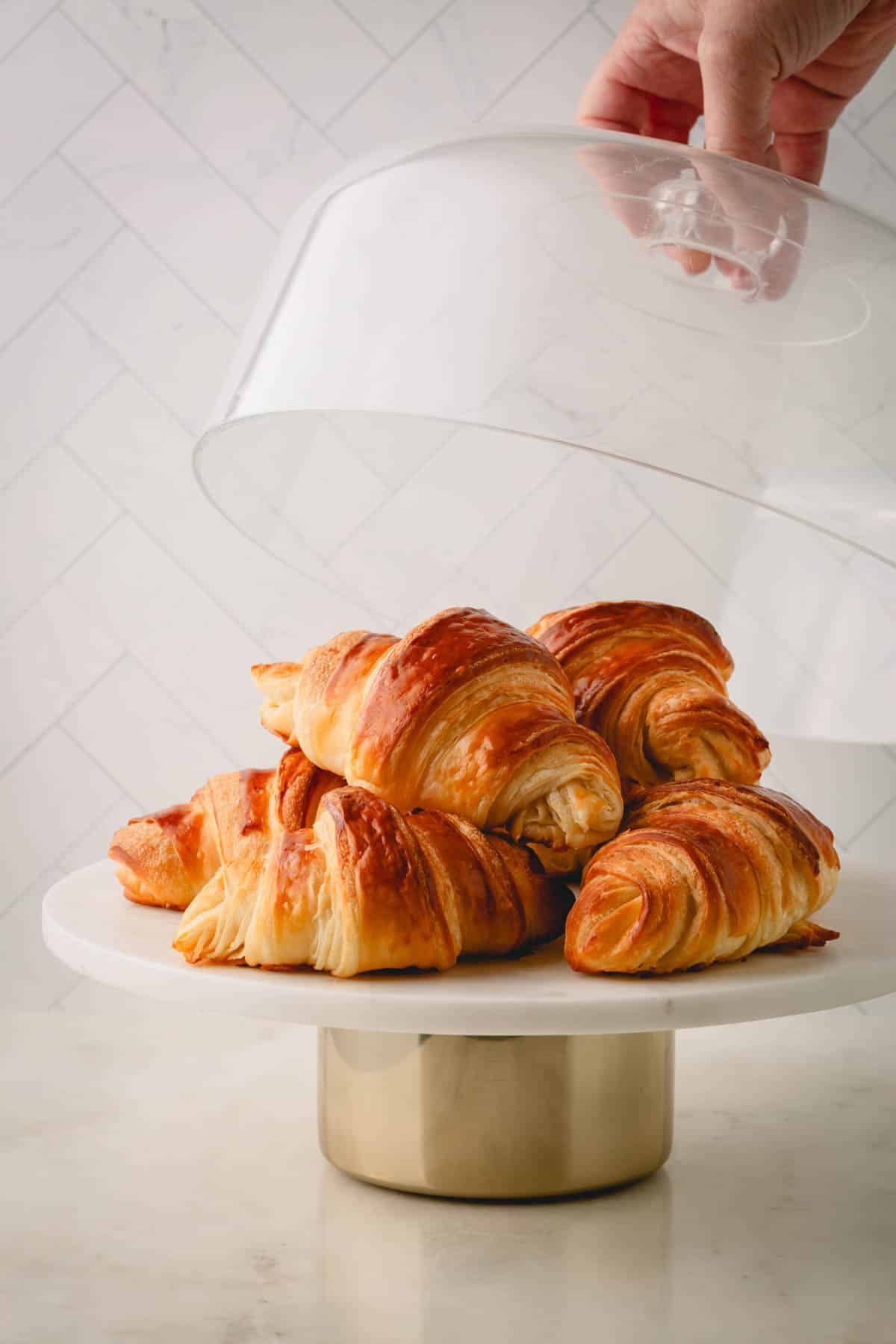 A white pedestal with homemade croissants and a hand covering them with a glass cover.