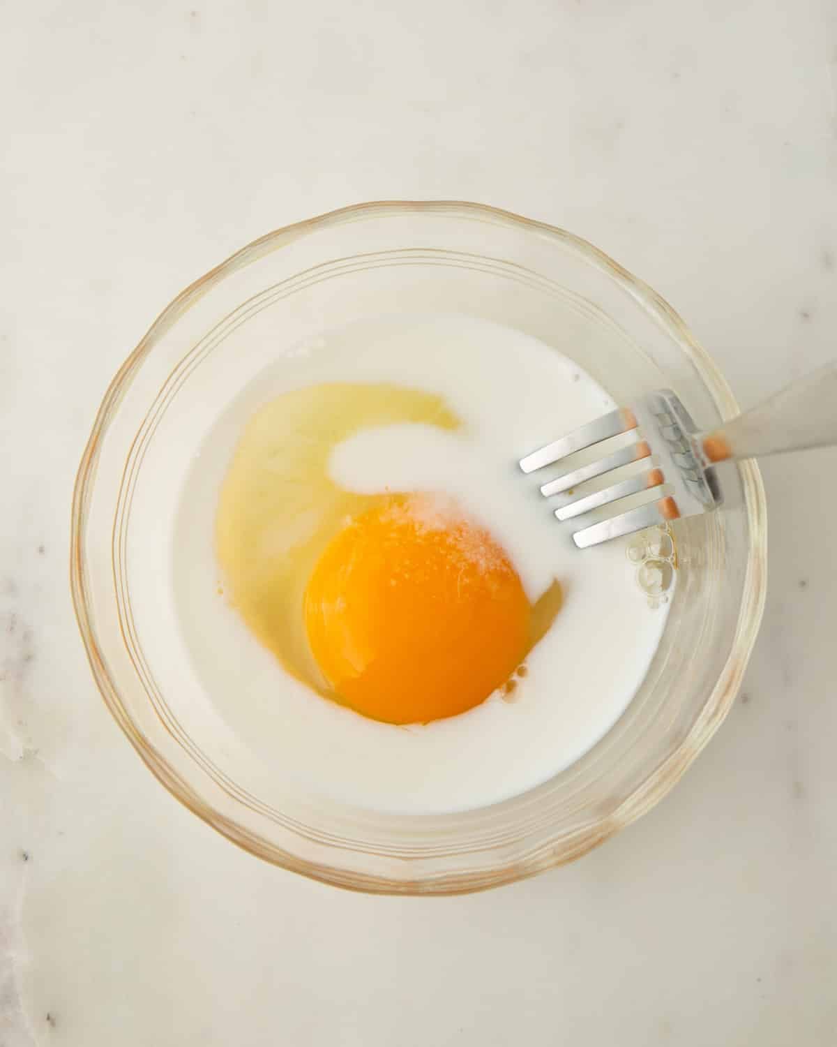 A glass bowl with egg and milk ready to mix with a fork.