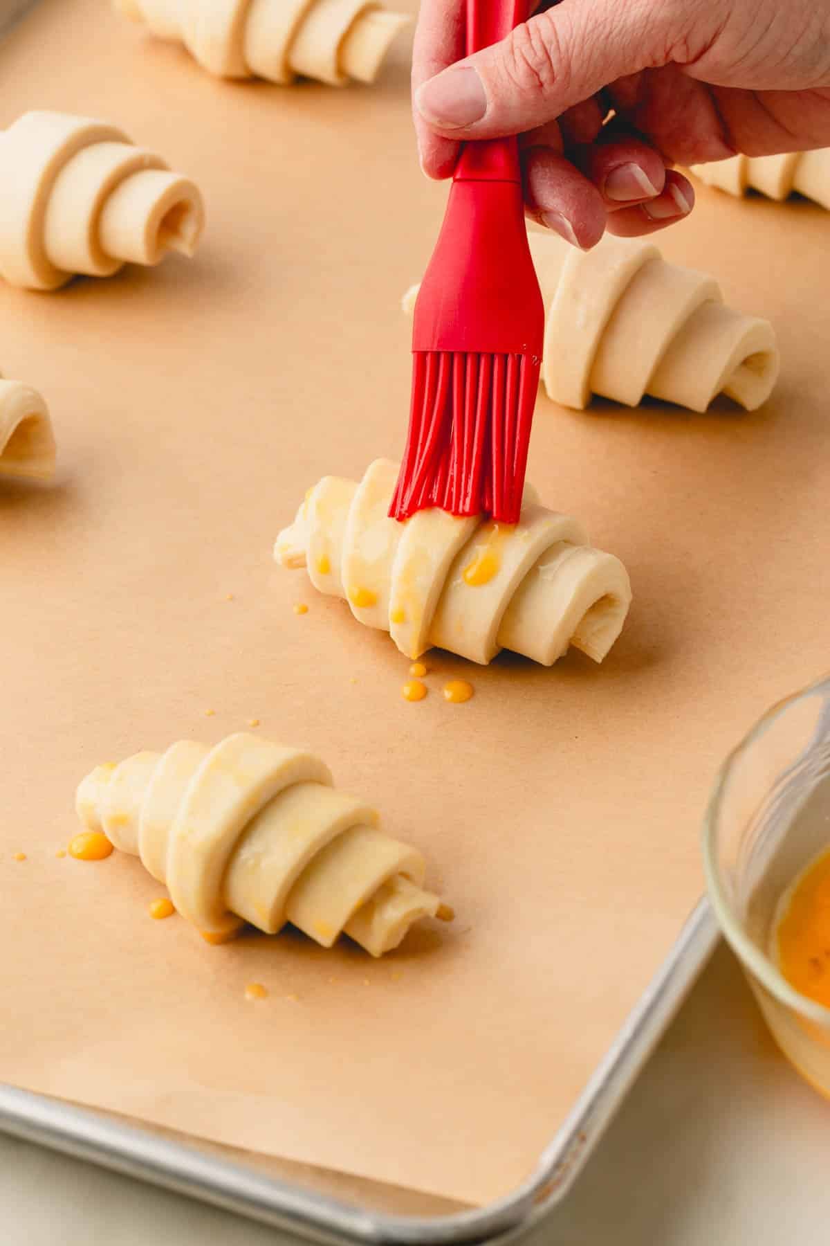 A red brush held by a hand brushing egg wash on rolled croissants.