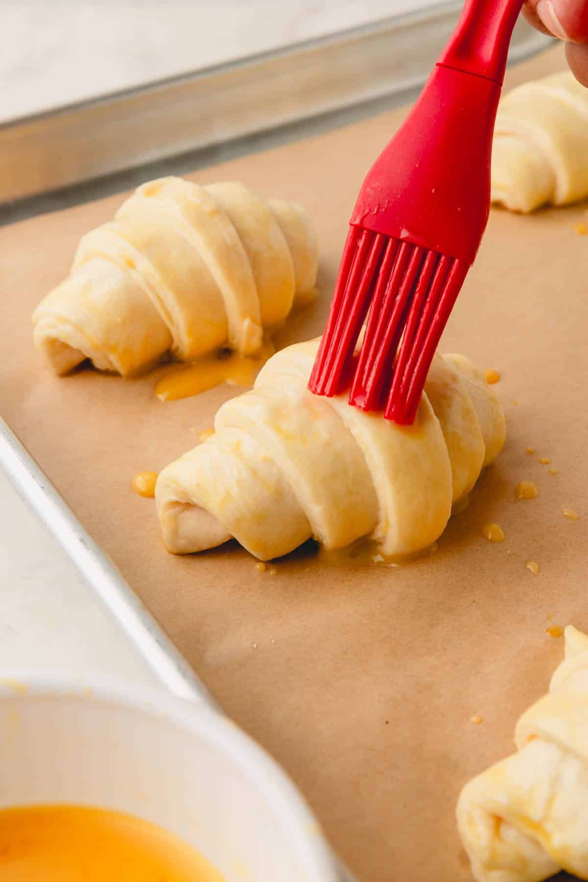 Close up image of red brush adding egg wash on rolled croissants.