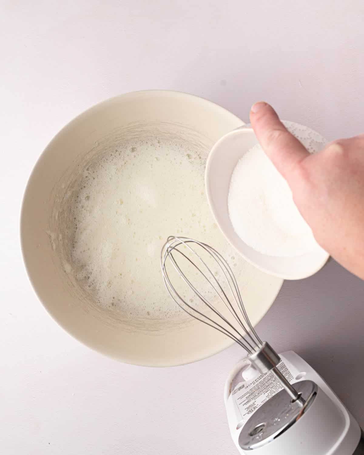 A white bowl with whipped egg whites and a hand adding a white bowl of sugar. A hand mixer is resting over the bowl.