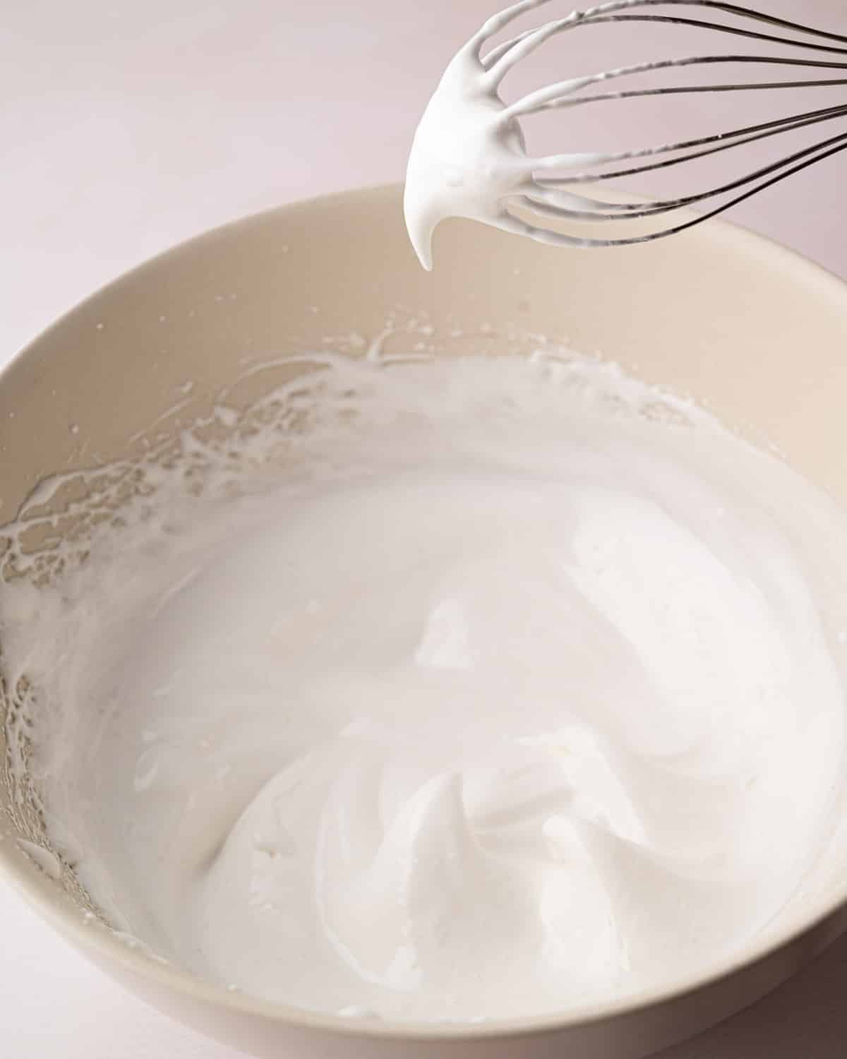 A white bowl with whipped egg whites with sugar and a hand mixer sitting on the side.