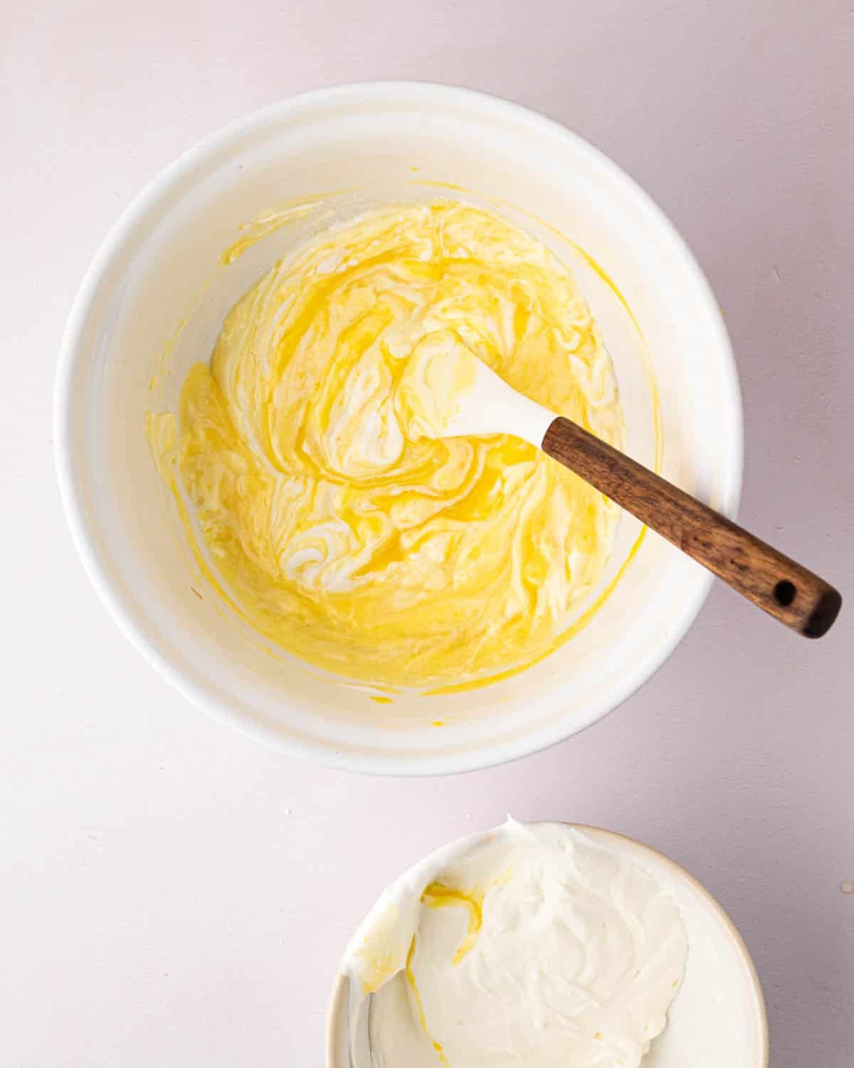 A white bowl with a white spatula and wooden handle resting on the side. The bowl has the thickened lemon base with whipped cream being folded in.