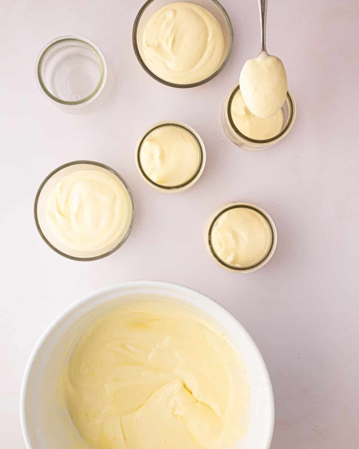 A white bowl with thickened lemon mousse. Small glass jars have lemon mousse scooped into them before chilling.