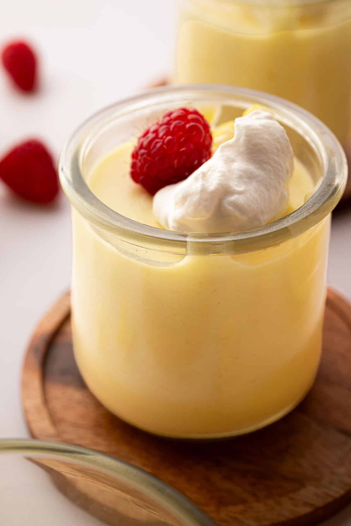 A small glass jar with lemon mousse and a raspberry on top. The glass jar is on a wooden coaster.