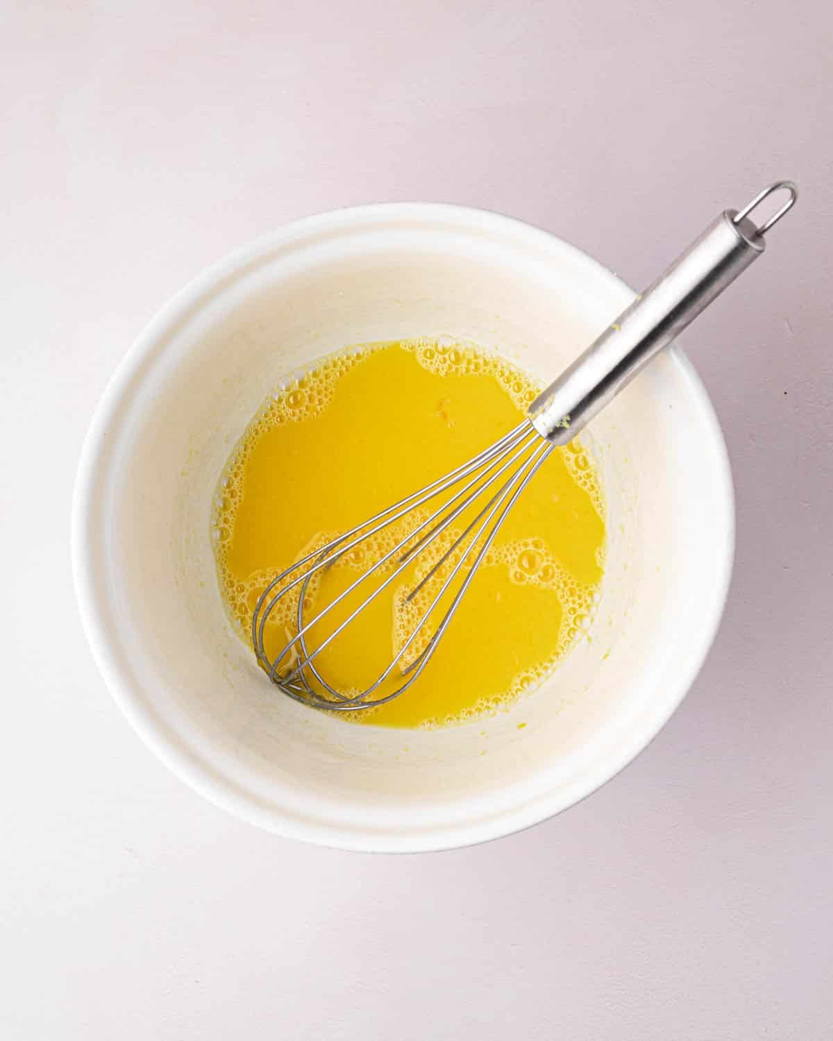 A white bowl with lemon juice and egg yolks with a whisk.