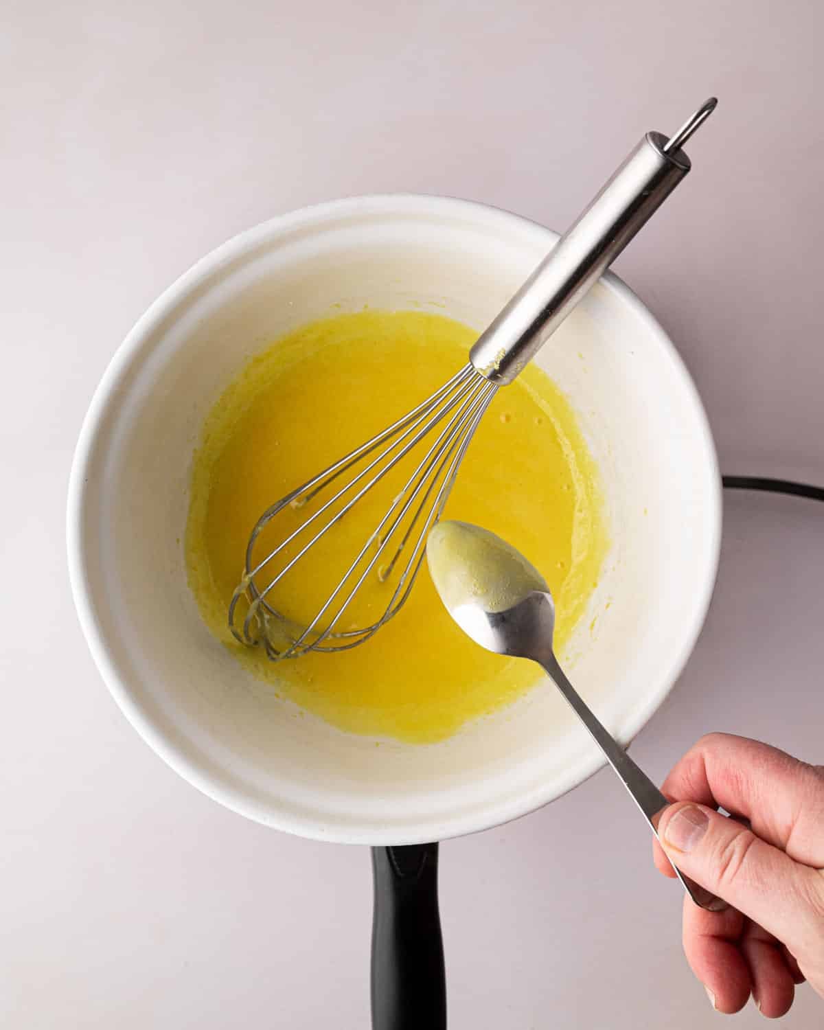 A white bowl over a double boiler with thickened egg yolks and lemon juice. A spoon is testing the thickness and a whisk is sitting on the side.