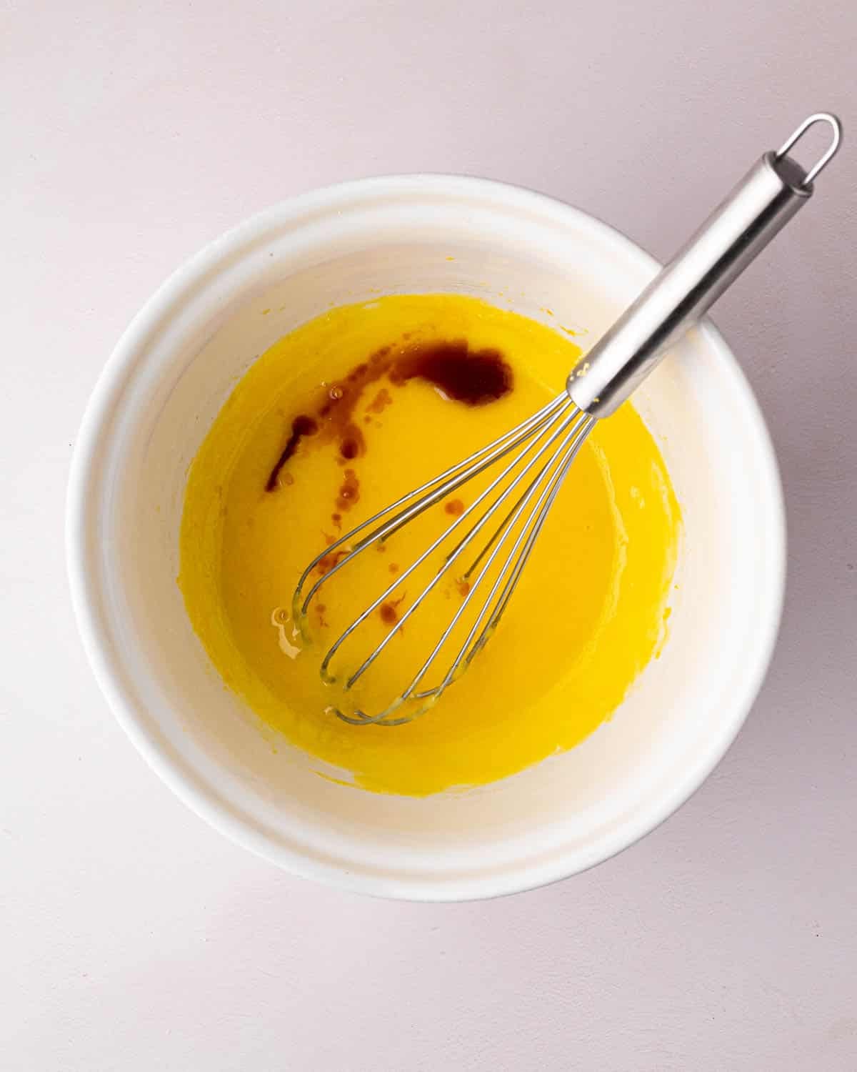 A white bowl with the cooked egg yolks and sugar and vanilla extract added. A whisk is resting on the side.