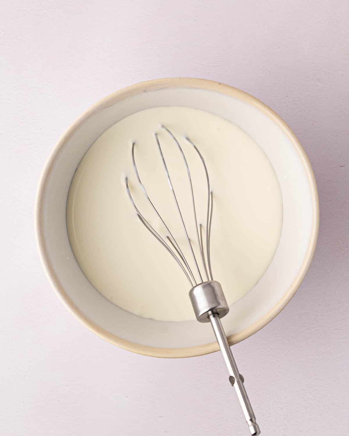 A white bowl with heavy cream ready to be whipped by the beater resting on the side.