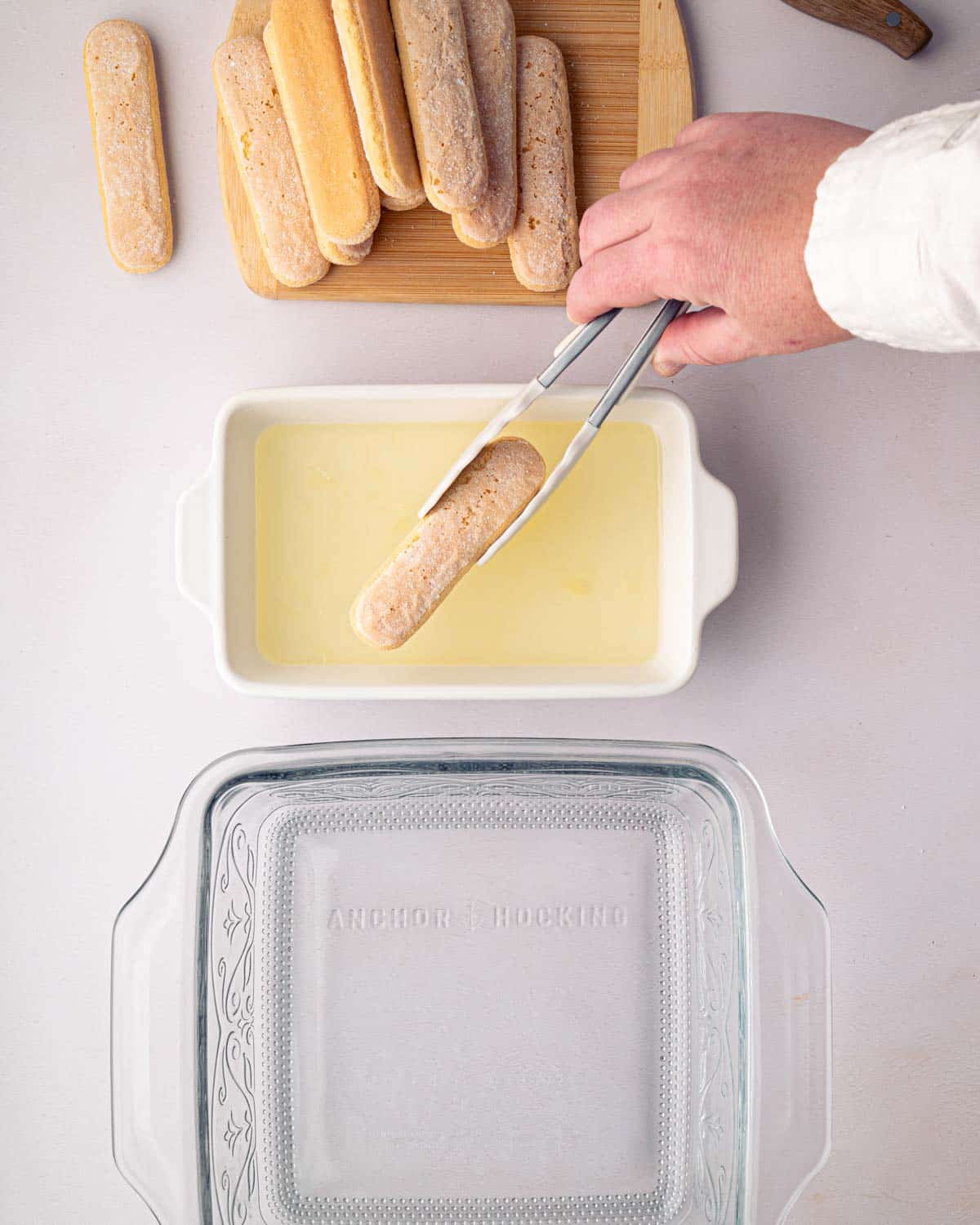 A hand dipping ladyfingers into lemoncello simple syrup in a small white baking dish with a glass baking dish on the side.
