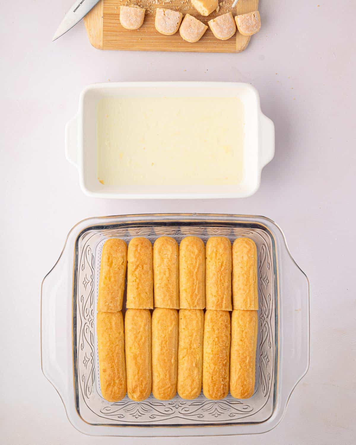 A white baking dish with lemon simple syrup is above a glass baking dish with dipped ladyfingers.