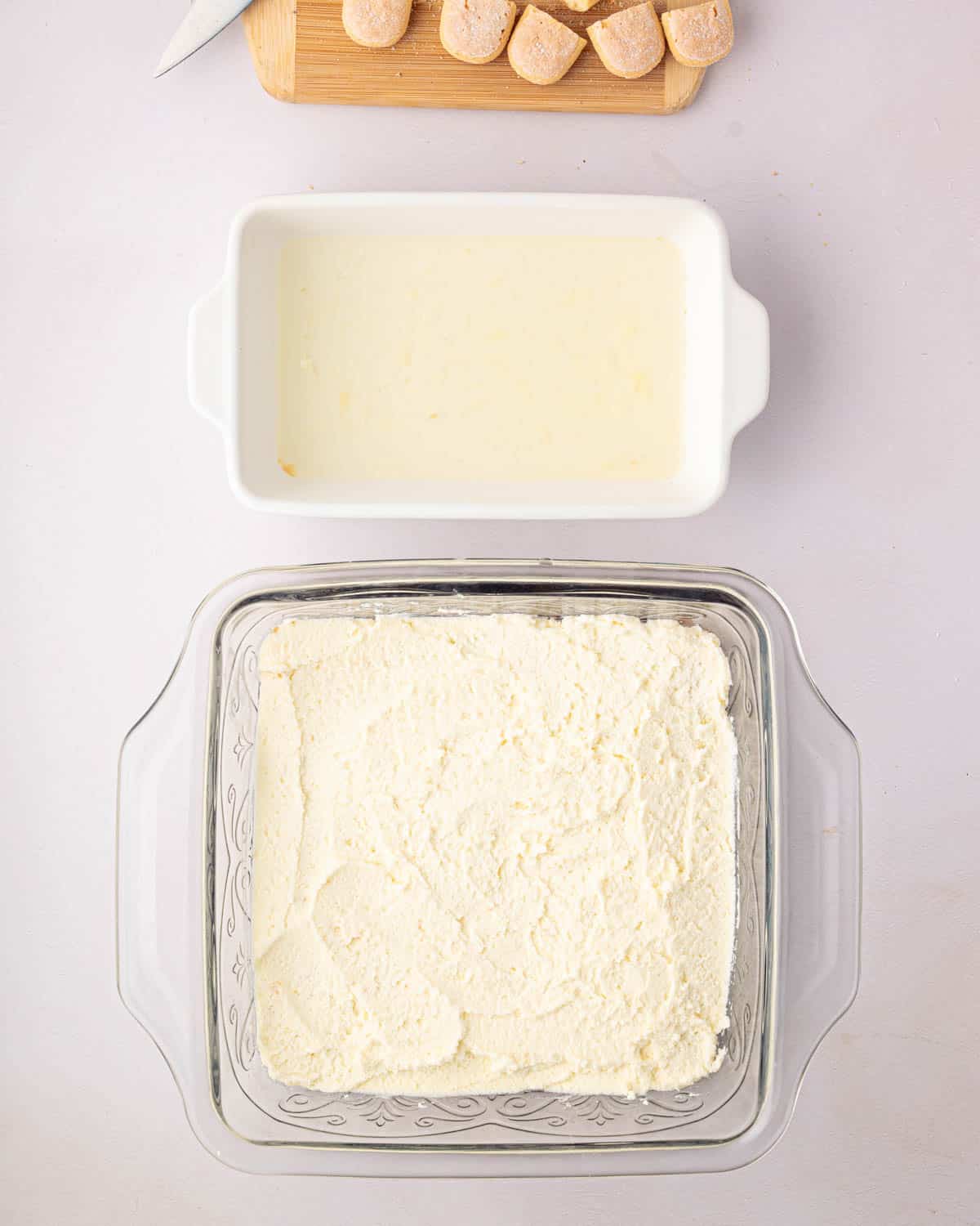 A white baking dish with lemon simple syrup is above a glass baking dish with dipped ladyfingers and a layer of mascarpone whipped cream.