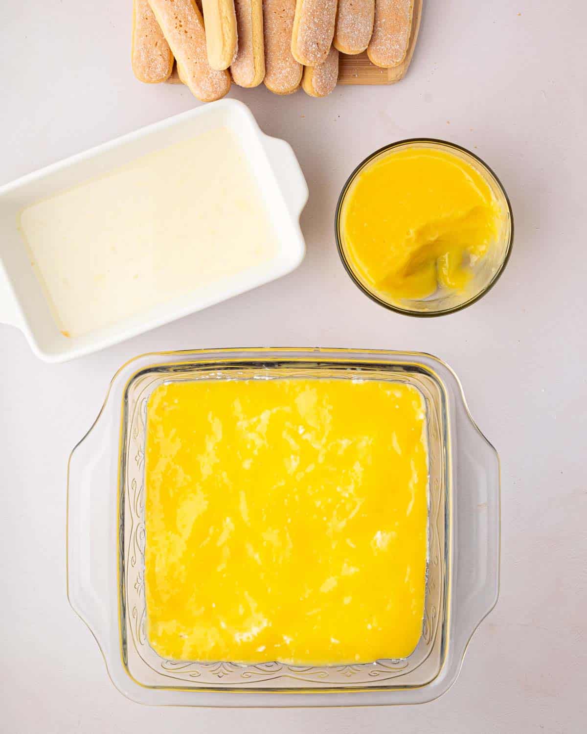 A glass baking dish has a layer of lemon curd for lemon tiramisu with a bowl of lemon curd to the side. A white baking dish with lemon simple syrup is above with ladyfingers on top of that.