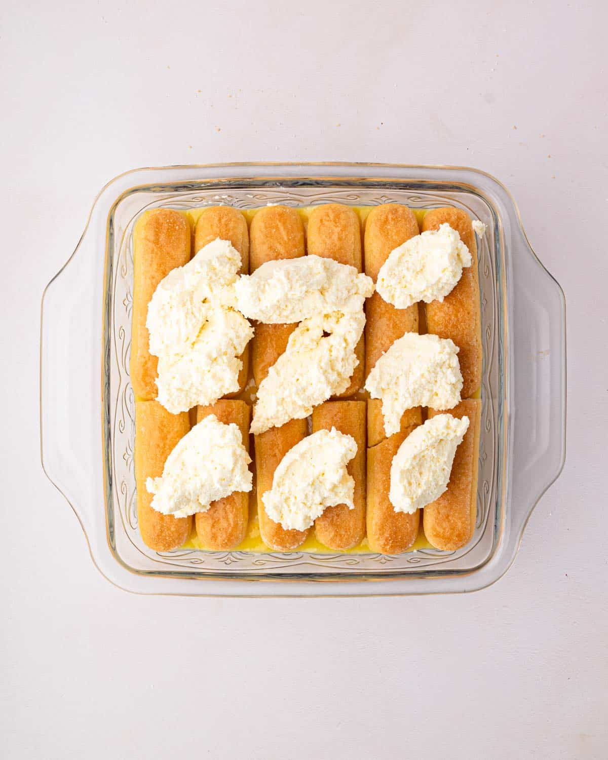 A glass baking dish with layers of lemon curd, mascarpone whipped cream, lady fingers, and dollops of mascarpone whipped cream.